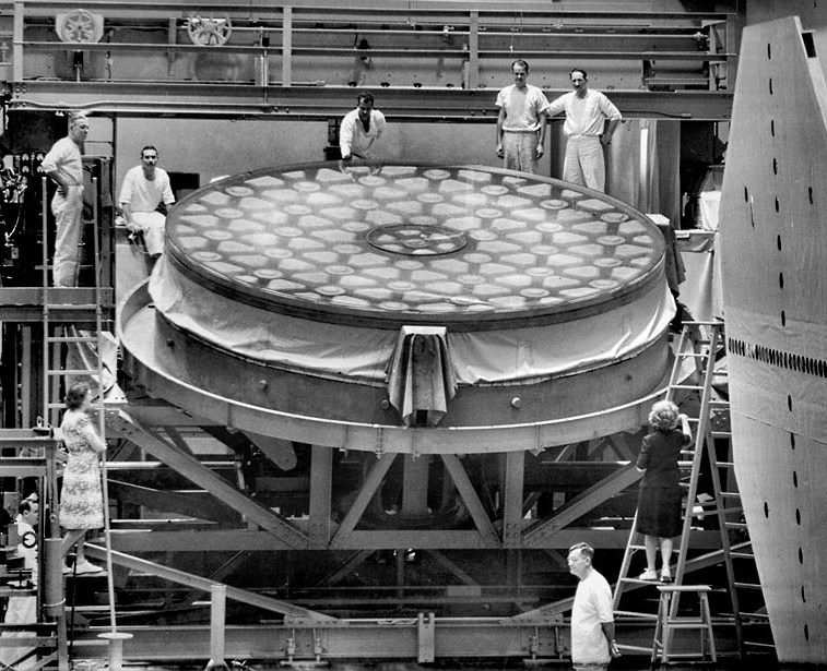 7 fair skinned men in their 30s or 40s stand around a large circular object, as well as two women on ladders on either side of the object. The object is round, large, and fabricated with mainly metal and fabric. The mirror of the object takes up the entire top surface, but it does not look like a mirror, that is to say it is not quite as reflective in this image as you would imagine a mirror to be. It looks to be almost a clear substance like acrylic and there are several uniform shapes within the acrylic: circles and triangles.