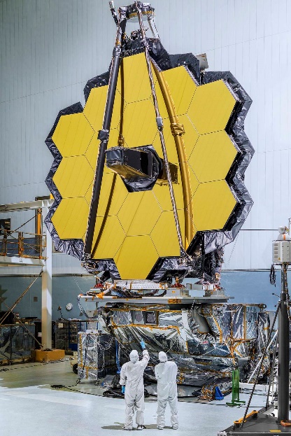 A large yellow structure stands above two workers that are covered head to toe in Personal Protective Equipment (PPE). The structure is in the shape of a honeycomb (a hexagon with 6 even sides) and is made up of several smaller honeycombs with an outline of a reflective material reminiscent of tin foil or mylar.