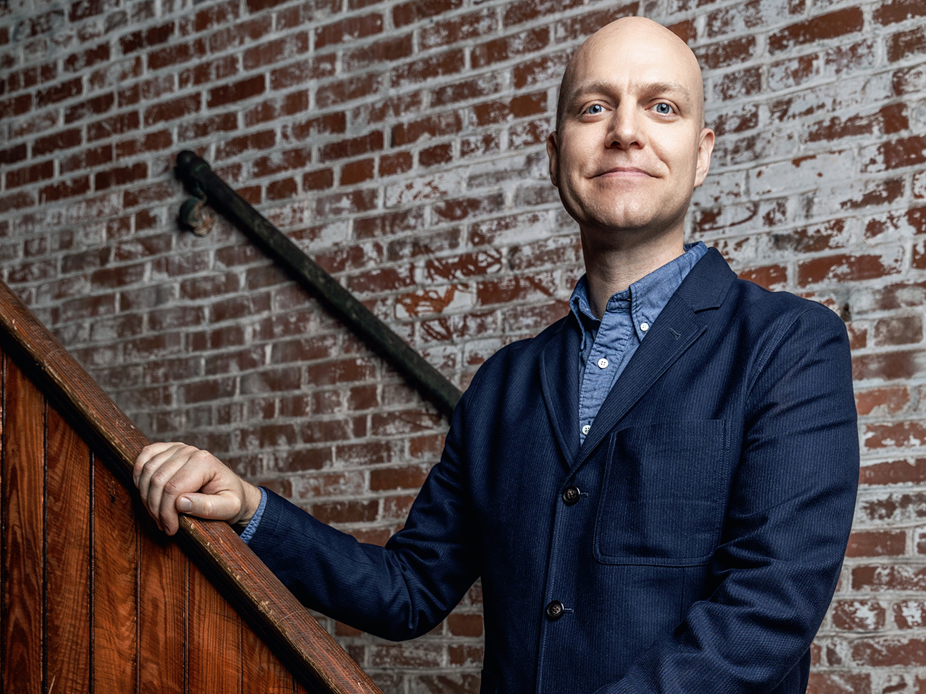 A bald man in a navy blazer stands confidently on a wooden staircase against a textured brick wall.