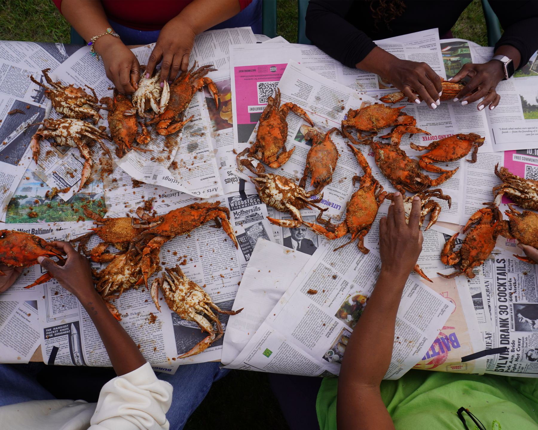 Twenty old bay encrusted bright orange and white crabs are splayed over black and white newspapers. Four sets of black hands are picking at the crabs.