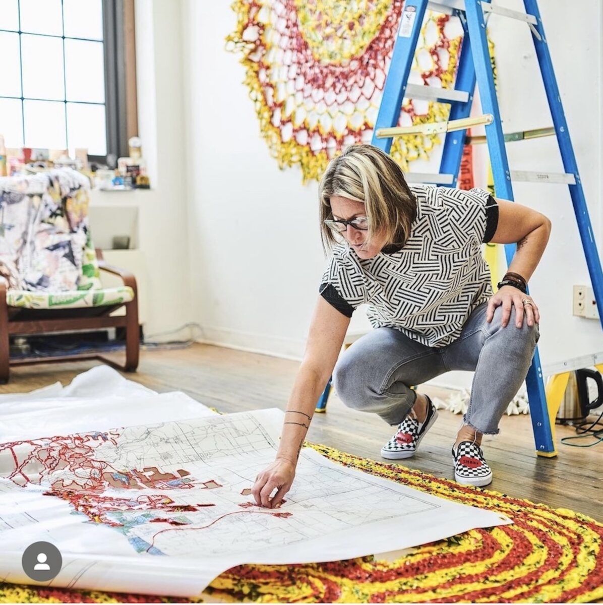 A person with short blonde hair is kneeling over their artwork on the floor as they work on it.
