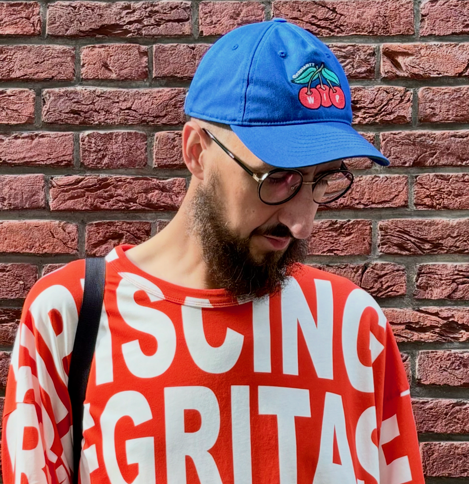 A bearded man in his forties is standing in front of a red brick wall. He looks down and is wearing a bright red t-shirt with large white letters, as well as a royal blue cap and glasses.