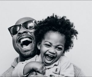 A close up of a man wearing sunglasses, smiling as he holds a smiling child at head level to him.