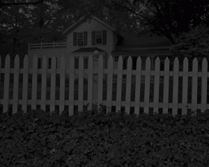 A white picket fence and white house are barely visible in the darkness of night.