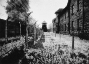 Behind the bars of a concentration camp. A wire fence stands on the left, and to the right, a brick building. In the center distance, a guard tower looms.