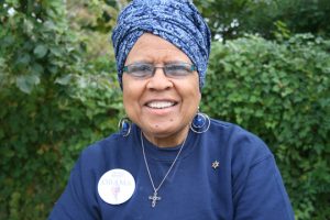 A portrait of a senior medium-dark skinned woman with a blue head wrap and blue glasses wearing a blue shirt, a large circular pin, a cross necklace, and hoop earrings. The woman is smiling and standing outside in front of greenery. 