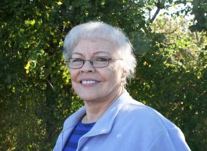 A portrait of a senior, light skinned woman with short white hair wearing thin glasses and a blue cardigan over a striped blue shirt. The woman is smiling and standing outside in front of greenery. 