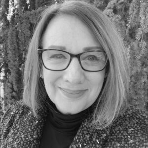 A black and white portrait of a smiling adult white woman with short brown hair and dark glasses. The woman is wearing a black turtleneck and a dark cardigan and is standing in front of a tree.