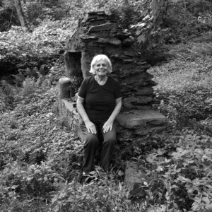A black and white image of a smiling white senior woman with short white hair who is wearing a dark short-sleeved shirt and dark pants. The woman is sitting outside on a large rock and is surrounded by trees and greenery. 