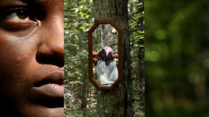 Close-up of half of a face with a dark brown skin tone and one eye gazing up is extremely close up on the left. On the right is a blurry green pixilated view of a wooded area. In the center is a mirror hanging on a tree in the forest. In the reflection of the mirror is a woman viewed from the back with a pink scarf over her long dark hair. Her medium brown arms reach up, and her hands disappear underneath her hair.