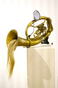 On a white art gallery pedestal, sits an assembled sculpture consisting of brass French horn, a bicycle mirror, and a black skateboard truck with two white wheels. A thick ponytail of blond hair emerges from just inside the bell of the horn and dangles out the entrance, handing down approximately one foot below.