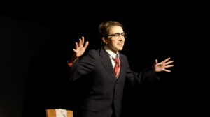 Light skinned, female-presenting figure in a men's suit, glasses, and a red tie stands beside in a dark space beside a wooden chair, both hands raised in an animated expression.
