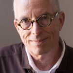 Marvin Heiferman, a man with a light complexion smiles and wears round-framed glasses and dark collared shirt layered over a white collared shirt and a white t-shirt.