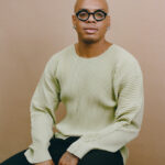 Meleke Glee, a man with a bald head, a thin moustache, light brown skin tone, and round, black-rimmed glasses wears a light sweater and sits on a stool and looks forward