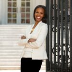 Sarah Lewis, a woman with medium brown skin tone, wears a white suit jacket and smiles with arms crossed as she leans against a wrought iron gate and looks forward.