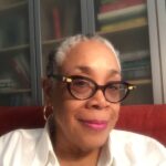 Lowery Stokes Sims, a woman with light brown skin tone wears glasses, a white shirt, golden earrings, and lipstick and smiles, looking forward at an angle