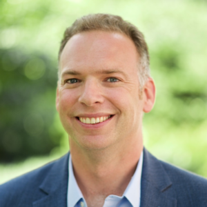 Matt Cregot, a man with a light skin tone and a navy blue suit jacket looks forward smiling